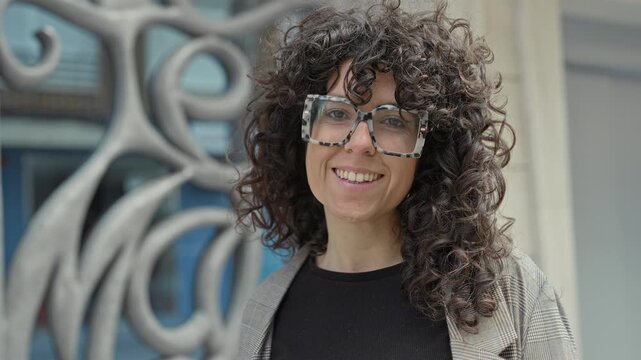 Woman wearing oversized tortoiseshell glasses smiles at camera in front of ornate building facade, curly hair visible; confidence authenticity.