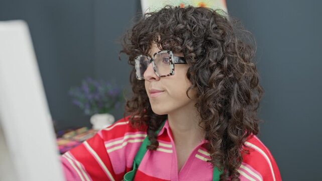 Woman artist paints canvas with brush at studio easel wearing apron and glasses; creative concentration.