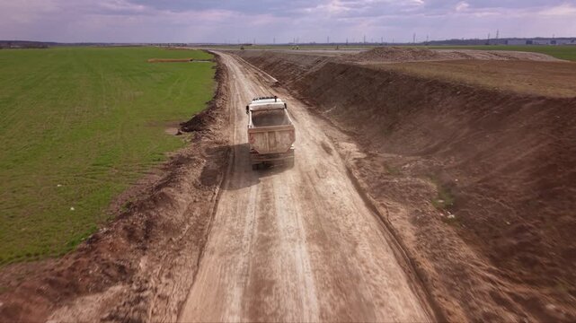 Aerial drone tracking shot of heavy dump truck transporting construction materials along temporary service road beside large highway expansion project with farmland, earthworks and infrastructure deve