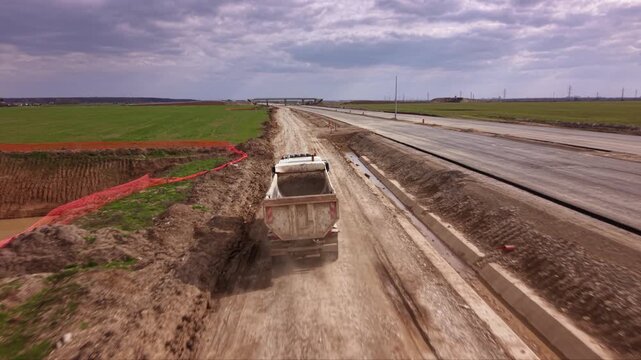 Aerial drone tracking shot of heavy dump truck transporting construction materials along temporary service road beside large highway expansion project with farmland, earthworks and infrastructure deve