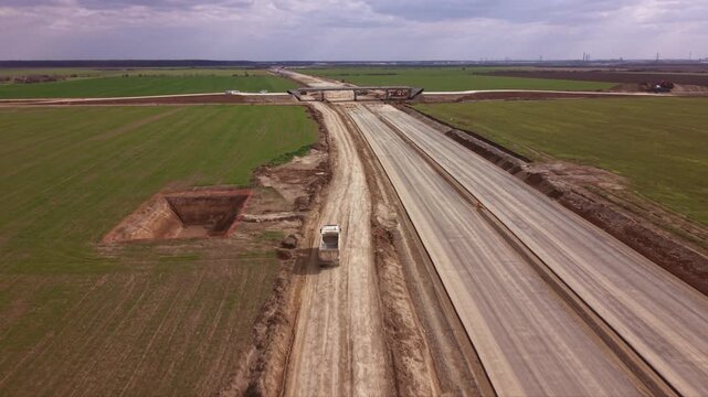 Aerial drone tracking shot of heavy dump truck transporting construction materials along temporary service road beside large highway expansion project with farmland, earthworks and infrastructure deve
