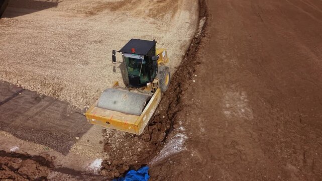 Aerial footage of a road roller compacting soil and aggregate during highway construction. The heavy compaction machine prepares the road base by compressing the ground, showcasing civil engineering, 