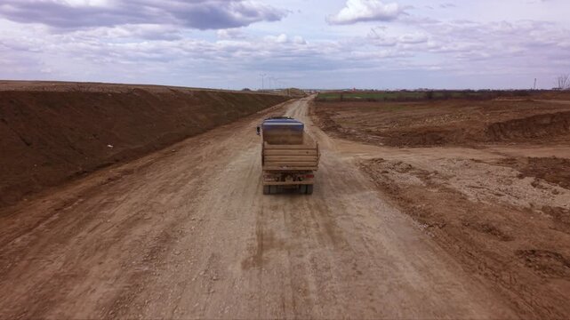 Aerial drone tracking shot of heavy dump truck transporting construction materials along temporary service road beside large highway expansion project with farmland, earthworks and infrastructure deve