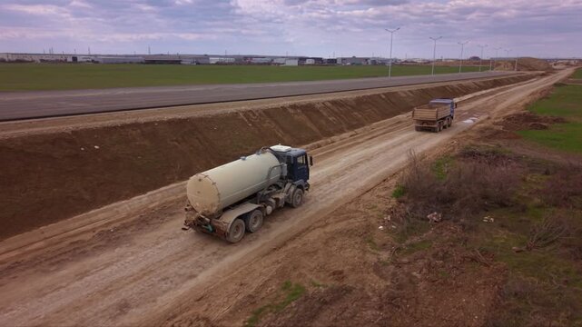 Aerial drone view of water tanker truck driving along unpaved road at large construction site, industrial vehicle supporting earthworks, road building, dust suppression and infrastructure development