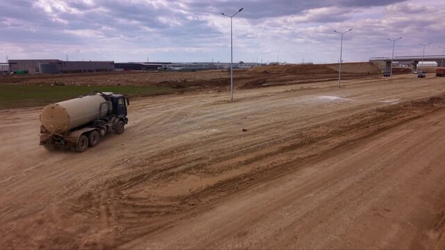 Aerial drone view of water tanker truck driving along unpaved road at large construction site, industrial vehicle supporting earthworks, road building, dust suppression and infrastructure development