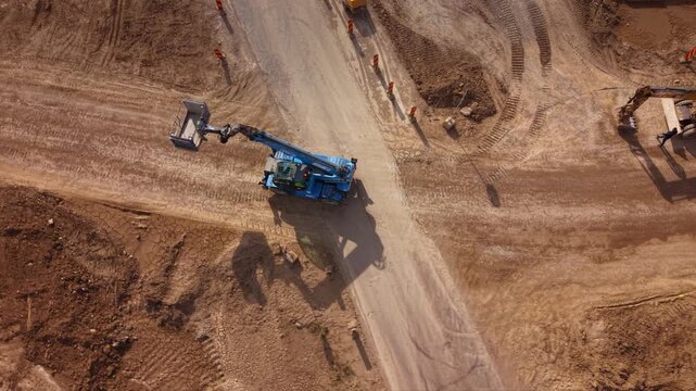 Aerial top down drone view of telehandler and hydraulic excavator working at road construction site with heavy machinery, earthmoving operations, dirt access roads and infrastructure development proje