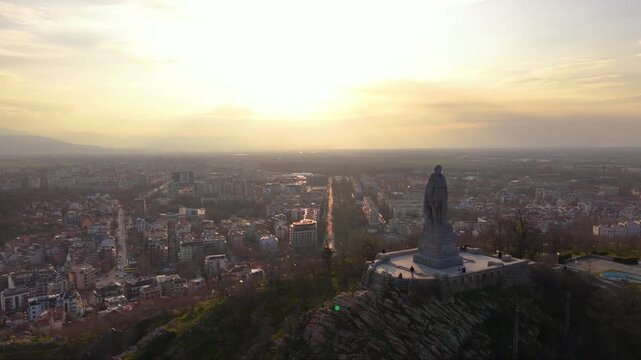 Aerial panoramic view of Alyosha Monument overlooking Plovdiv city at golden hour with sprawling urban skyline, Rhodope Mountains in the distance, historic landmark and dramatic evening clouds, Bulgar