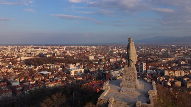 Aerial panoramic view of Alyosha Monument overlooking Plovdiv city at golden hour with sprawling urban skyline, Rhodope Mountains in the distance, historic landmark and dramatic evening clouds, Bulgar