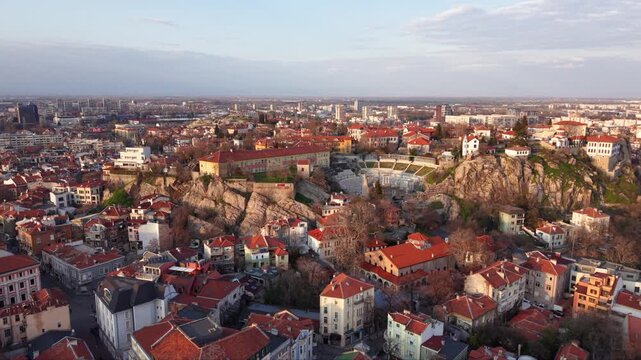 Aerial panoramic view of Plovdiv city skyline at golden hour featuring Nebet Tepe hill, historic Old Town, dense urban residential districts and modern cityscape under dramatic spring clouds, Bulgaria