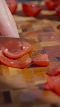 Hand slices a small tomato with knife on wooden cutting board, with cut pieces nearby. Concept of fresh ingredients and recipe preparation