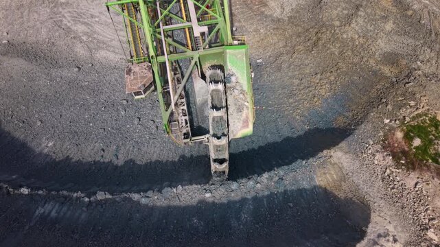 Aerial footage of a giant bucket wheel excavator continuously digging and loading rock in an active open pit quarry. The enormous mining machine transfers excavated material onto conveyor belts, showc