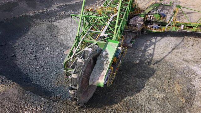 Aerial footage of a giant bucket wheel excavator continuously digging and loading rock in an active open pit quarry. The enormous mining machine transfers excavated material onto conveyor belts, showc