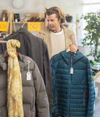 Curly haired man stands near the rail with outerwear and chooses a raincoat and a down jacket. Shopper at a clothing sale in a store © JackF