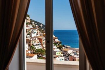 Obraz premium View from open window to colorful buildings on seaside hill and blue ocean. Daytime sunny weather offers bright coastal landscape. Curtains frame scenic Mediterranean town.