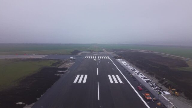 Aerial view straight down an airport runway disappearing into dense fog, with fresh runway markings, threshold stripes and surrounding construction areas creating a dramatic low-visibility aviation sc