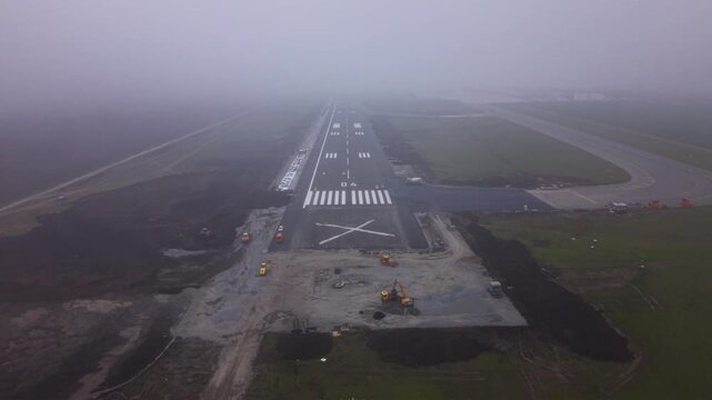 Aerial view straight down an airport runway disappearing into dense fog, with fresh runway markings, threshold stripes and surrounding construction areas creating a dramatic low-visibility aviation sc