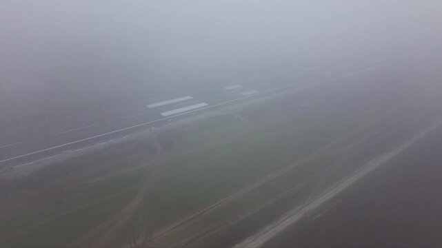 Aerial view of an airport runway almost completely obscured by dense fog, with only faint runway threshold markings and pavement visible through extremely low-visibility weather conditions.