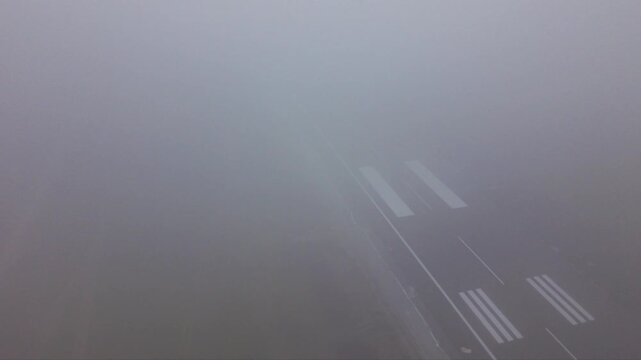 Aerial view of an airport runway almost completely obscured by dense fog, with only faint runway threshold markings and pavement visible through extremely low-visibility weather conditions.