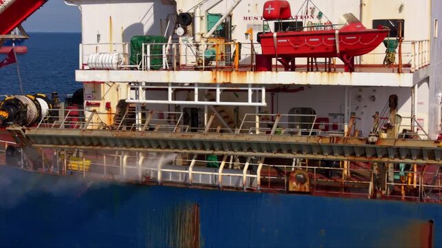 Close aerial view of a large commercial vessel with crew members working along the deck beside a weathered blue hull, showing lifeboats, railings, marine equipment and signs of rust and heavy maritime