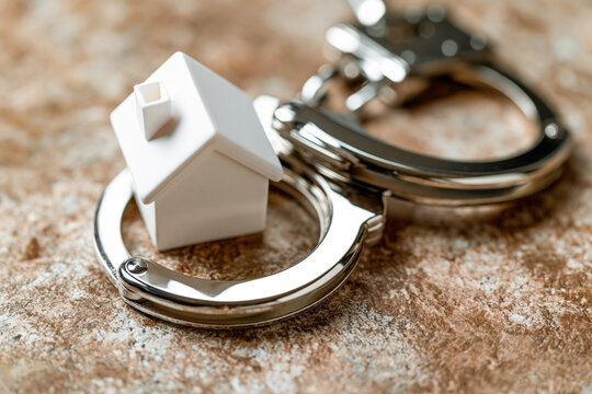 A small white toy house placed inside shiny metal handcuffs on a textured surface. Conceptual image representing mortgage debt, real estate fraud, or property arrest