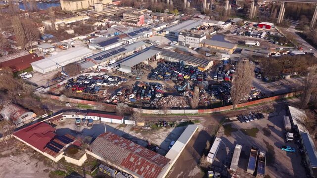 Top-down aerial view of a large vehicle scrapyard packed with rows of dismantled cars, scrap metal and automotive parts beside industrial buildings, showing vehicle recycling and waste processing.