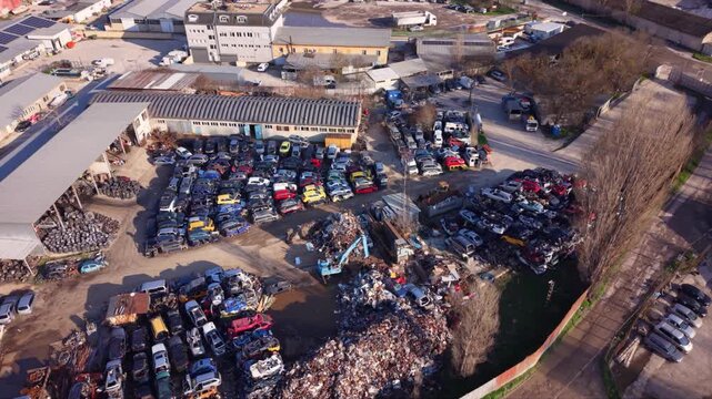 High-resolution aerial drone footage of an automotive scrapyard featuring hundreds of abandoned vehicles, scrap metal piles, industrial warehouses, and a vehicle recycling facility. Top-down view high