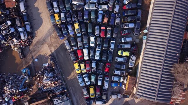 High-resolution aerial drone footage of an automotive scrapyard featuring hundreds of abandoned vehicles, scrap metal piles, industrial warehouses, and a vehicle recycling facility. Top-down view high