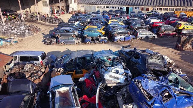 Top-down aerial view of a large vehicle scrapyard packed with rows of dismantled cars, scrap metal and automotive parts beside industrial buildings, showing vehicle recycling and waste processing.