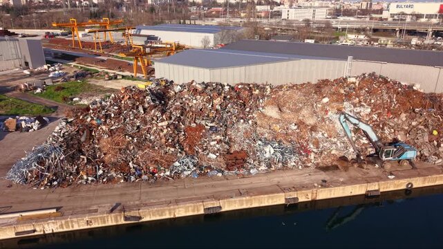 Aerial footage of a large scrap metal recycling facility at an industrial port, featuring massive stockpiles of ferrous and non-ferrous metal ready for processing. Warehouses, cranes and waterfront in