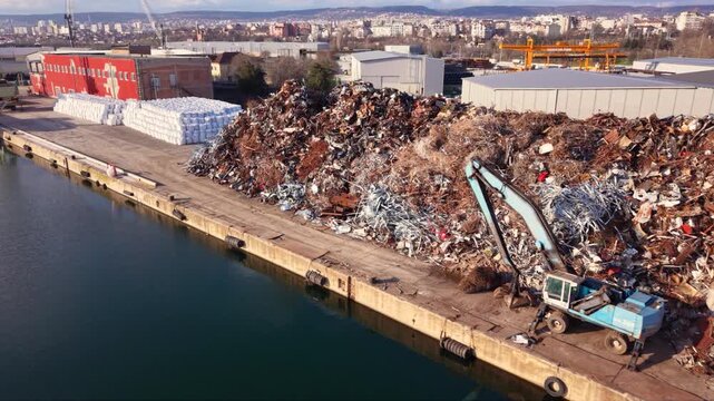 Aerial footage of a large scrap metal recycling facility at an industrial port, featuring massive stockpiles of ferrous and non-ferrous metal ready for processing. Warehouses, cranes and waterfront in
