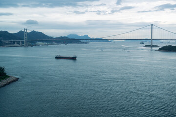 Fototapeta premium Large cargo ship sailing under suspension bridge on calm water during cloudy day with distant mountains