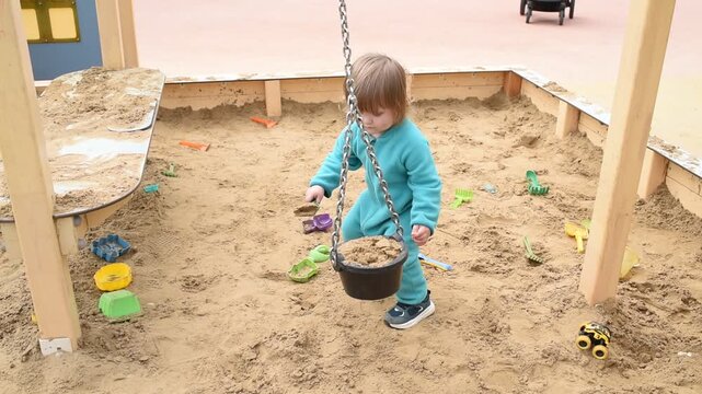 Toddler boy scooping sand and pouring into hanging bucket on playground 