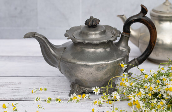 Two old metal teapots are sitting on a wooden table. Tiles are visible in the background. Chamomile flowers are lying next to the teapots.