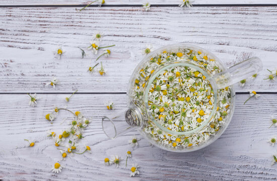 A glass teapot sits on a light-colored wooden surface. The teapot is filled with chamomile flowers. The teapot is photographed from above.