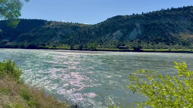 Beautiful green fast moving Montana river with solitary cabins on the opposite bank. There are beautiful mountains in background. 