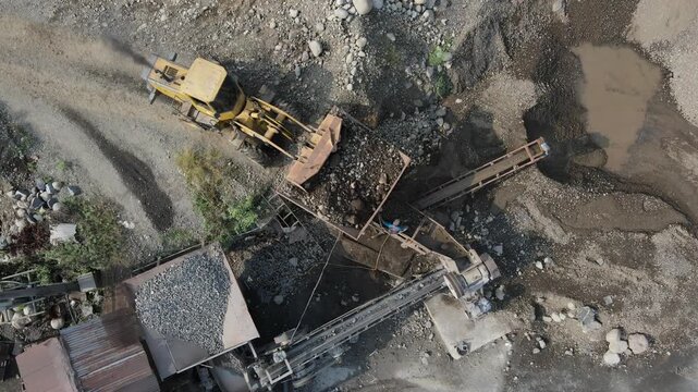 Aerial view of a yellow bulldozer loading rocks into a stone crusher at a construction site with conveyor belts in Patikraja, Central Java, Indonesia.
