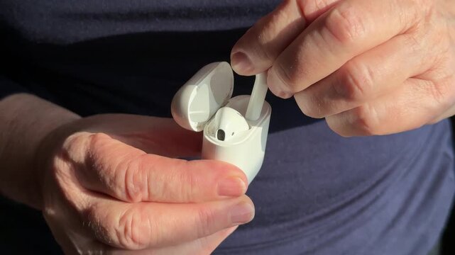 Closeup woman holding white earbud case, gently removing single earbud for listening warm sunlight highlights textured hands