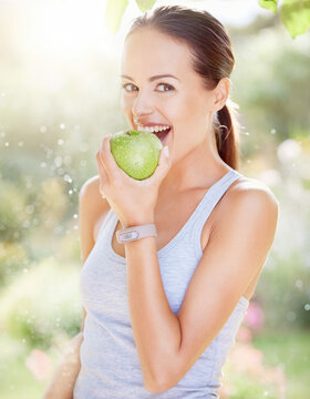 Young woman eating a fresh green apple outdoors. She wears a fitness tracker on her wrist and grey tank top, enjoying a healthy snack in a sunlit setting.