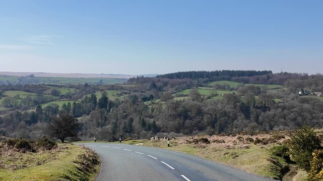 Dartmoor Devon England UK.  27.03.2026. Video. A paved road curves through a picturesque landscape of green fields,  on Dartmoor Devon with dense forests, and rolling hills under a bright blue sky