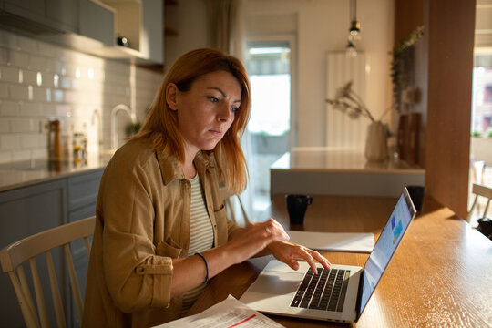 Focused woman paying bills on laptop at home kitchen