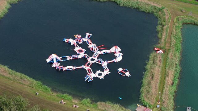 Aerial view of Fenland Aqua Park featuring colorful inflatable obstacles on a lake surrounded by green reeds and solar panels in Whittlesey, England, United Kingdom.