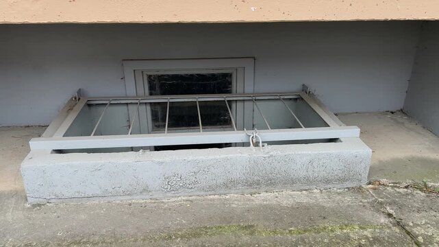 Metal grating on the basement window, concrete ledge under the balcony of a high-rise building, spring daylight, weathered paint, moss on the asphalt, chips