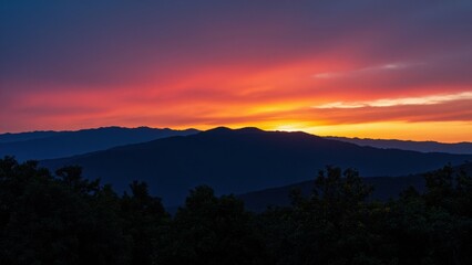 Fototapeta premium Mountain Serenity at Dusk: A breathtaking vista of silhouetted mountains unfolds under a vibrant sky painted with hues of deep purple, fiery orange, and soft yellow as the sun dips below the horizon.