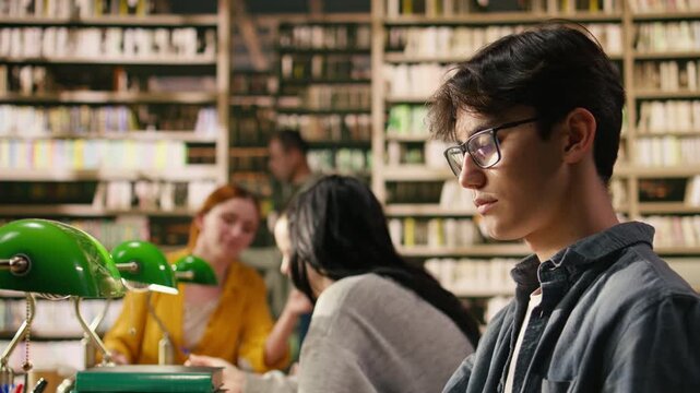Portrait of young man reading open book at library study table for self study session, creating academic scene with stacked books near green lamps and bookshelves, Classmates study indoors.