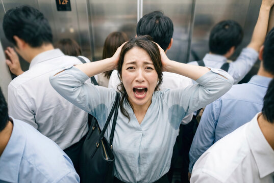 満員エレベーターで脇汗対策の制汗スプレーを使うのを忘れたことに気づき嘆いている、多汗症のシャツで汗ジミが目立つ日本人女性会社員