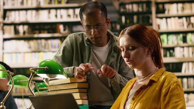 Tutor explaining assignment to student and pointing at laptop during study session, mentoring young adult in finishing research.