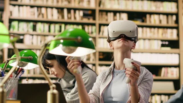 Student wears VR glasses and holds controllers in university library, using immersive technology beside classmates with open books, Virtual reality technology in education, digital learning.