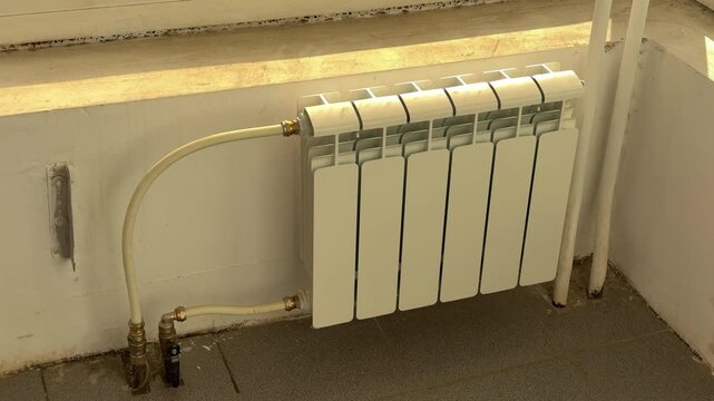 White panel radiator appears mounted beneath stone sill in practical heating room, showing pipe connections, brass valve fittings, wall brackets, chipped plaster, floor tiles, and close