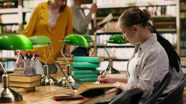 Student in university library distracted by smartphone notification, pauses writing assignment and spends time on phone, Closing laptop and books and texting.