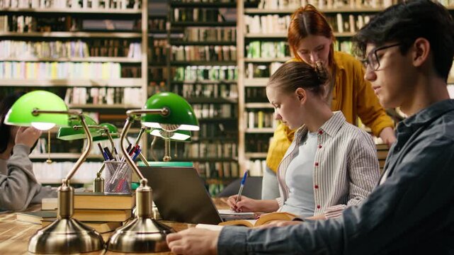 Tutor and student discuss research beside laptop and stacked books in library, offering academic guidance with collaborative learning at study desk with green.